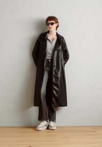 Fekete műbőr trench coat laza fazonnal, szürke gombos ing, szürke farmernadrág és fehér vastag talpú sneaker. A napszemüveg kontrasztot ad.