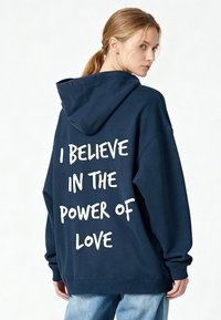 Sweat à capuche bleu marine avec une coupe décontractée, présentant un grand texte blanc au dos qui dit "JE CROIS AU POUVOIR DE L'AMOUR."