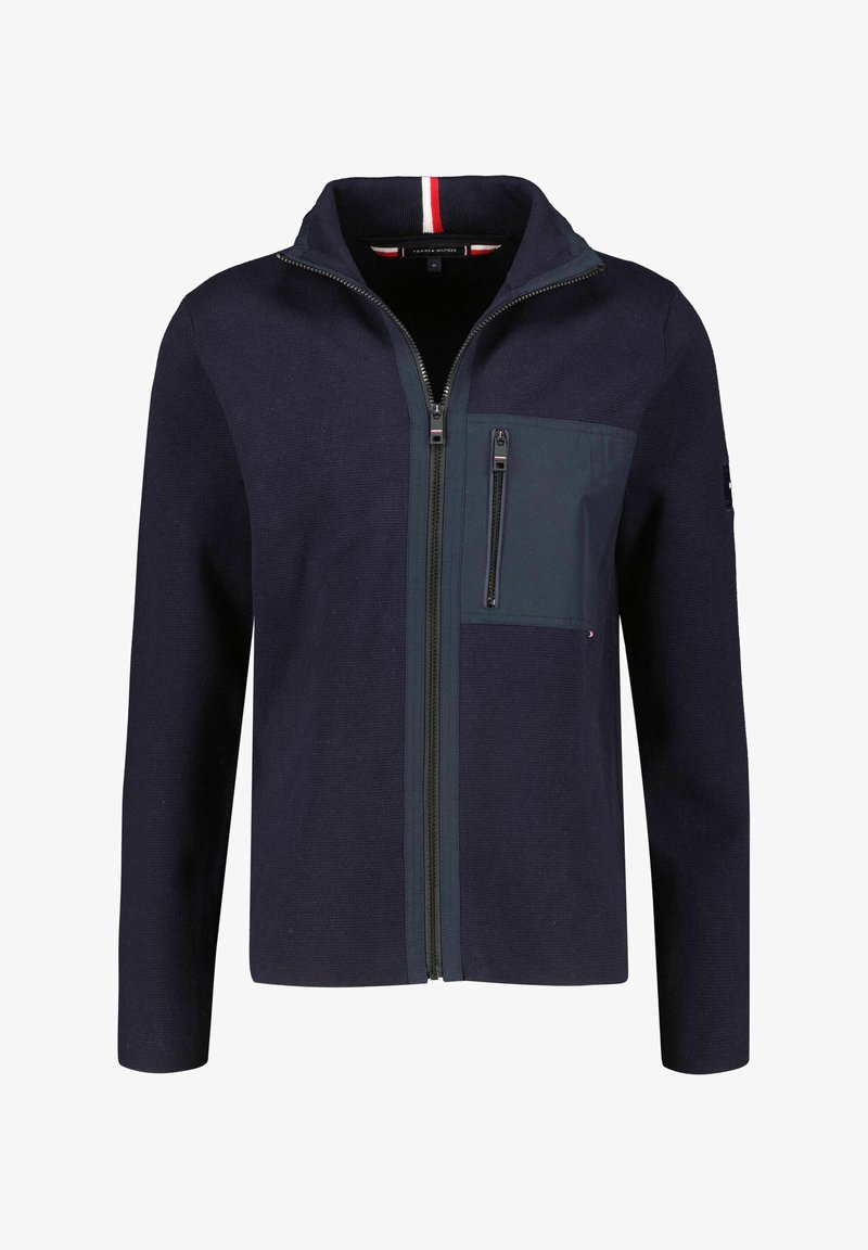 Veste zippée bleu marine avec col montant, poche zippée sur la poitrine et bord rouge, blanc et bleu subtil à l'intérieur du col.