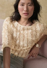 Top beige à sequins avec un design texturé et des franges, à manches courtes et encolure ronde, associé à un jean en denim clair.