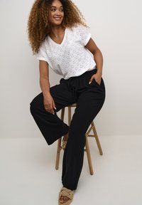 Witte katoenen top met decoratieve gaten, gecombineerd met zwarte wijde broek van een zachte stof. Sandalen met een geknoopte, gestructureerde stijl.