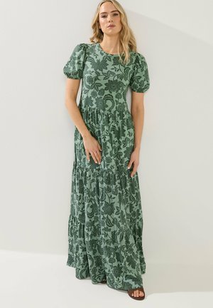 Nainen pukeutunut vihreään maksi-mekkoon, jossa tumma kukkakuosi ja liehutetut lyhyet hihat, seisoo vaalean tasaisen seinän edessä.