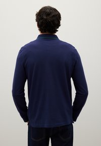 Mann mit dunklem, lockigem Haar, der ein navyblaues langärmliges Poloshirt und dunkle Jeans trägt und mit den Händen in den Taschen vor einem schlichten Hintergrund steht.