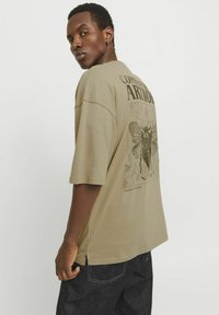 Camiseta oversize verde oliva con mangas cortas, que presenta un gráfico negro de una polilla en la parte posterior y aberturas en los lados del dobladillo.