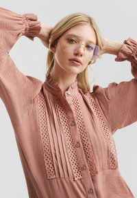 Blouse rose pâle avec un tissu texturé, col volanté, boutons à l'avant et détails en dentelle le long de la patte de boutonnage. Manches longues à volants.