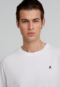 Witte katoenen t-shirt met een ronde halslijn, voorzien van een klein marineblauw doodskop- en kruisbeenderenlogo op de linkerkant van de borst. Soepele textuur.