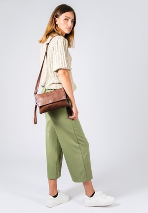 Borsa a tracolla in pelle marrone con texture morbida, forma rettangolare, chiusura con zip e tracolla regolabile. L'outfit include un top in maglia beige e pantaloni verdi.