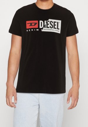 Mann trägt ein schwarzes Diesel T-Shirt mit rotem und weißem Logo und hellblauen Jeans vor einfarbigem Hintergrund.