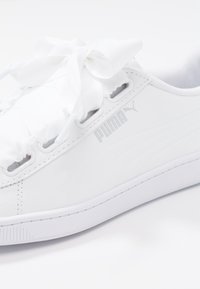 Sneakers bianchi Puma con superiore in materiale sintetico liscio, lacci testurizzati morbidi, dettaglio logo argento e suola piatta in gomma.