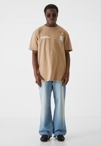 Brown oversized cotton t-shirt with front text graphic, paired with light blue flared jeans and black sneakers. Smooth texture, relaxed fit.