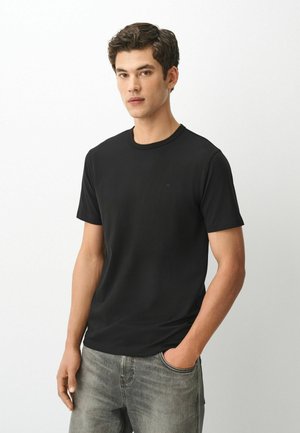 Ung mand med kort mørkt hår iført en enkel sort kortærmet t-shirt og grå jeans, med den ene hånd i lommen, stående mod en hvid baggrund.