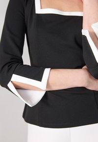 Haut noir avec un décolleté carré, une bordure blanche et des manches trois-quarts avec des fentes ouvertes aux poignets. Tissu lisse et texturé.