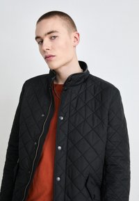 Μαύρο ραφτό μπουφάν με ψηλό γιακά, που διαθέτει κουμπιά με κλιπ και φερμουάρ. Φοράει ένα πουκάμισο χρώματος σκουριάς με υφή.