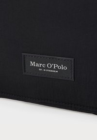 Sac noir en tissu texturé avec un bord cousu. Arborant une étiquette noire avec un texte blanc : "Marc O'Polo EST. À STOCKHOLM."