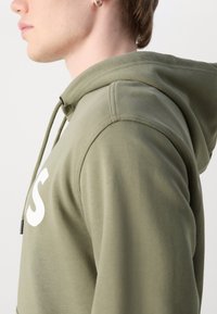 Jeune personne portant un sweat à capuche vert olive avec texte blanc, montrant un profil de côté et une épaule, sur un fond clair uni.