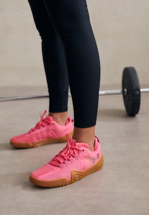 Persoon met zwarte leggings en felroze sportschoenen die bij een halter met gewichten op een gymvloer staat.