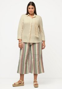 Beige skjorte med knapper og en tekstureret makramé lomme med frynser, parret med stribede vide bukser i grønne, lilla og creme farver.