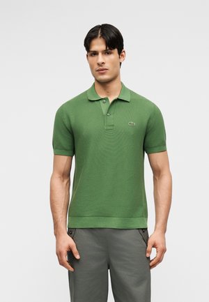 Homme debout portant un polo vert à manches courtes et un pantalon gris, sur un fond uni gris clair.