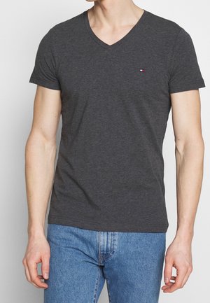 Mann trägt dunkelgraues V-Ausschnitt-T-Shirt mit kurzen Ärmeln und blaue Jeans, steht mit entspannten Armen an den Seiten.
