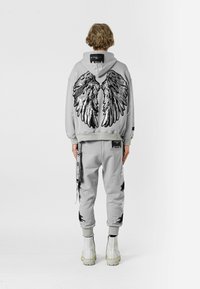 Sudadera gris con grandes gráficos de alas en negro y plata en la parte posterior, combinada con joggers a juego con detalles desgastados y acentos en negro.