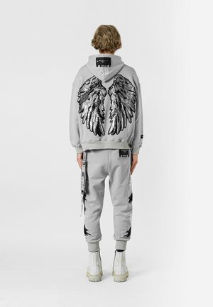 Sudadera gris con grandes gráficos de alas en negro y plata en la parte posterior, combinada con joggers a juego con detalles desgastados y acentos en negro.