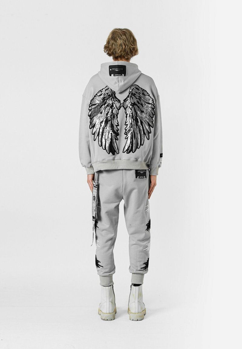 Sudadera gris con grandes gráficos de alas en negro y plata en la parte posterior, combinada con joggers a juego con detalles desgastados y acentos en negro.
