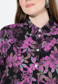 Camicia nera a fiori con bottoni, caratterizzata da grandi fiori rosa e verdi, colletto classico e bottoni in plastica. Il tessuto ha un aspetto testurizzato.