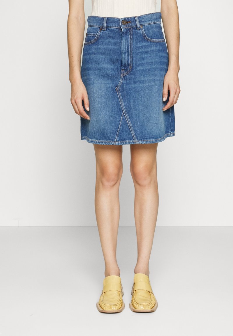 Femme portant une jupe en denim bleue longueur genou et des chaussures jaunes sans lacets, debout devant un fond clair uni.