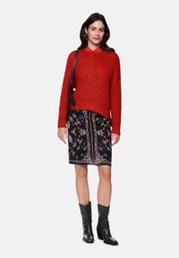 Red knitted sweater with a loose fit, paired with a black skirt featuring floral patterns and a decorative stripe; black heeled boots.