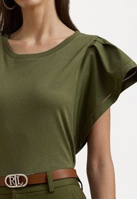 Oliivinvihreä lyhythihainen paita, jossa on rypytetty hartiadesign, pyöreä kaula-aukko ja herkät aukko-osoitukset, yhdistettynä ruskeaan vyöhön, jossa on hopeinen solki.