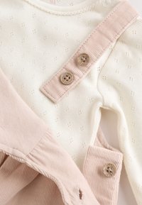 White baby outfit with small eyelet pattern and pink corduroy straps fastened by round brown buttons labeled "NEXT."