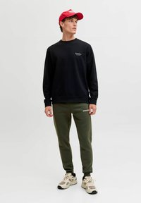 Sort sweatshirt med broderet logo, olivengrøn tapered sweatpants, hvide sneakers med beige detaljer og en rød kasket.
