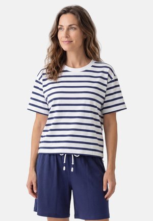 Kobieta w białej i granatowej koszulce w paski z krótkim rękawem oraz granatowych szortach ze sznurkiem, stojąca na jednolitym tle.