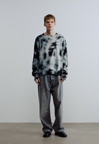 Φούτερ με ασπρόμαυρο σχέδιο tie-dye και στρογγυλή λαιμόκοψη, συνδυασμένο με φαρδιά γκρι τζιν και μαύρα παπούτσια. Ουδέτερο φόντο.