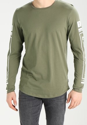 Homme portant une chemise à manches longues vert olive avec texte graphique blanc sur les manches et un jean gris foncé, debout devant un fond uni.
