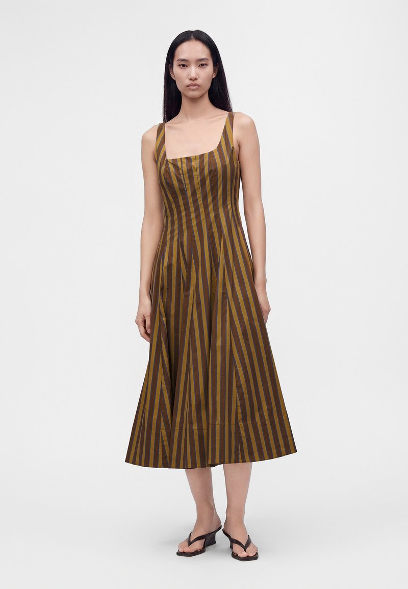 Femme portant une robe midi sans manches à rayures verticales marron et moutarde avec un col carré et des sandales noires à talons, debout sur fond blanc.