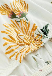 Blomstret stof med gule og lyserøde blomster på en hvid baggrund, med samlet elastikdetaljering langs kanterne. Blød tekstur.