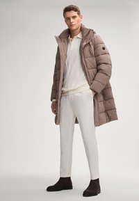 Bruine pufferjas met een hoge kraag en rits aan de voorkant, gedragen over een lichtcrème gestructureerd langemouwen top en witte broek, gecombineerd met donkerbruine laarzen.