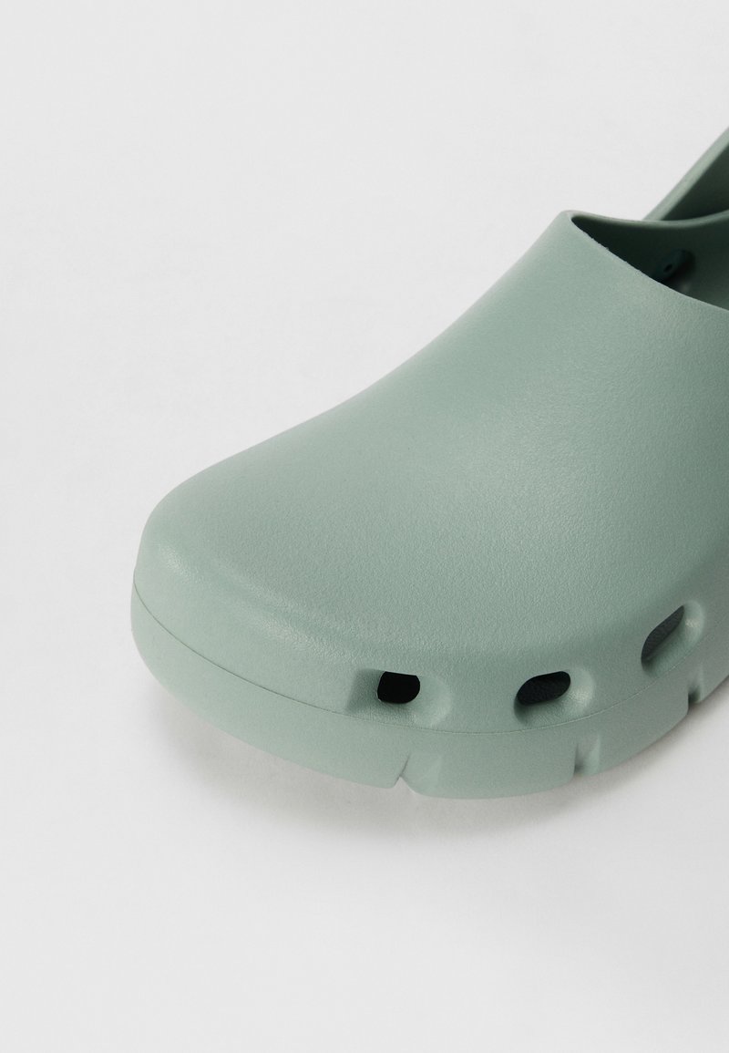 Zoccoli verde chiaro realizzati in materiale liscio e flessibile con bordi arrotondati. Presentano fori di ventilazione e un design a punta chiusa.