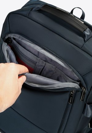 Zaino blu con una superficie testurizzata, dotato di un compartimento con zip aperto che rivela un rivestimento grigio e tasche in rete rossa all'interno.