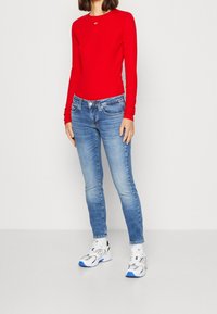 Blusa roja de manga larga combinada con vaqueros ajustados de color azul claro. El conjunto incluye zapatillas blancas con acentos plateados y detalles azules.