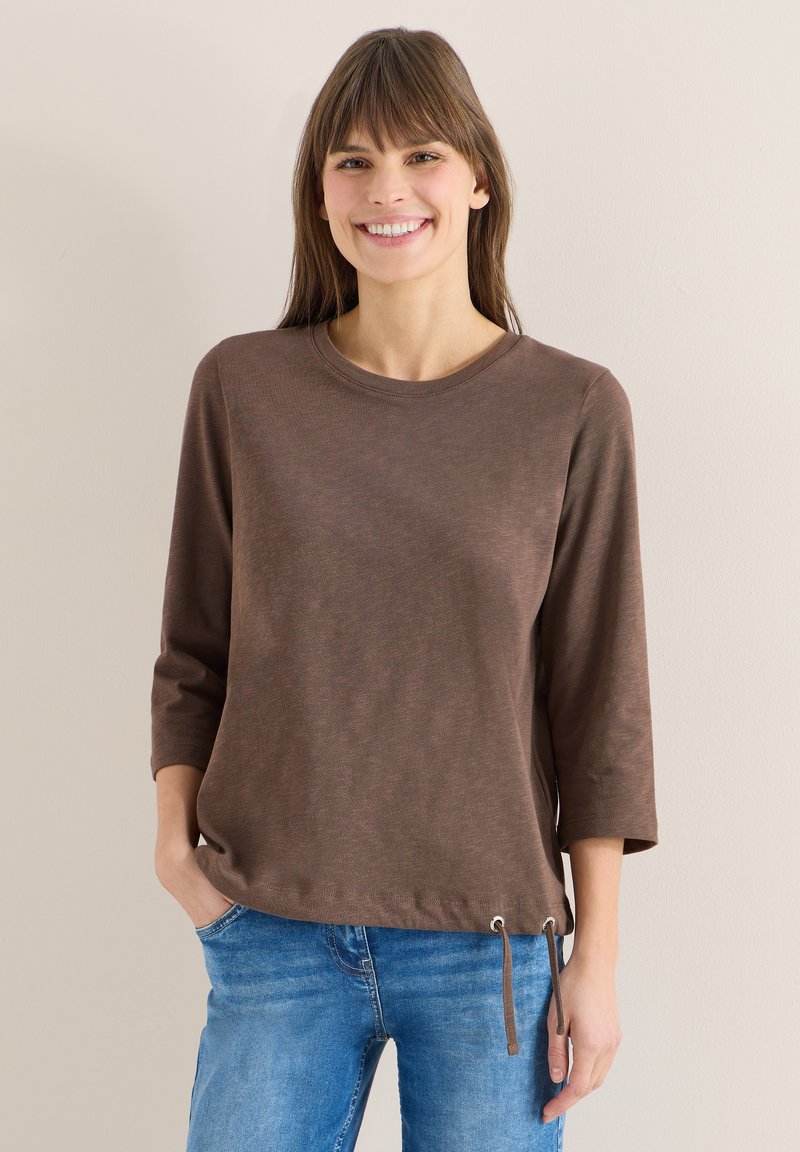 Femme souriante, portant un haut marron à manches longues avec des cordons au niveau de l'ourlet et un jean bleu, debout devant un fond clair uni.