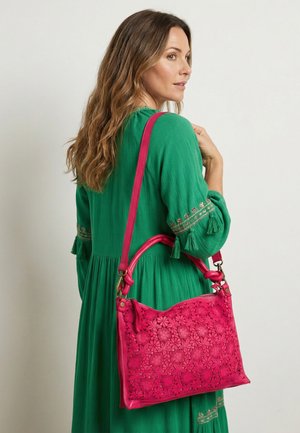 Woman in green dress carrying bright pink floral-patterned handbag over her shoulder, turned slightly to the side against plain background.