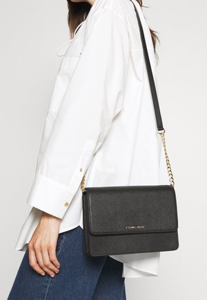 Frau in weißem Hemd und blauer Jeans trägt eine schwarze Michael Kors Umhängetasche mit goldener Kettenriemen auf ihrer Schulter.