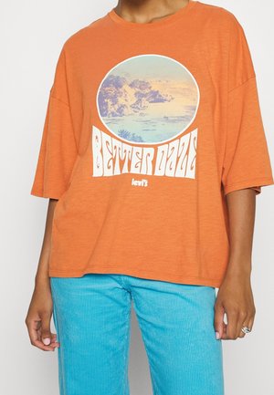 Personne portant un t-shirt orange Levi’s avec une impression circulaire d’un paysage et le texte "Better Days", associé à un pantalon en velours côtelé bleu clair.