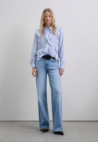 Woman with long blonde hair wearing a light blue ruffled blouse, wide-leg light blue jeans, black belt, and black pointed shoes, standing on gray floor.