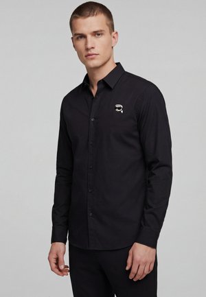 Giovane uomo che indossa una camicia nera abbottonata con un piccolo logo grafico di un volto con occhiali da sole sul petto sinistro, in piedi davanti a uno sfondo grigio uniforme.