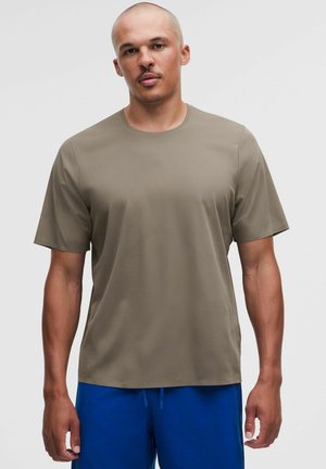 Mann mit rasiertem Kopf, der ein schlichtes olivgrünes T-Shirt mit kurzen Ärmeln und leuchtend blaue Shorts trägt und vor einem weißen Hintergrund steht.