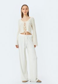 Witte cropped trui met gestructureerde brei en een strikdetail aan de voorkant, gecombineerd met losse, witte, gestructureerde broek en beige instappers.