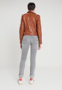 Person med kort mörkt hår som bär en brun läderjacka, ljusgrå uppvikta jeans och vita sneakers med röda hälflikar som står vänd bort.